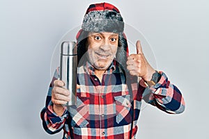 Handsome mature handyman wearing winter hat with ear flaps holding thermo smiling happy and positive, thumb up doing excellent and