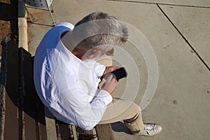 Handsome mature man with beard and grey hair is sitting thoughtfully on a park bench while consulting his mobile phone. The man is