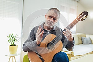 Mature man in casual clothes is smiling while playing guitar at home