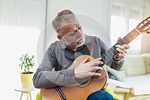 Mature man in casual clothes is smiling while playing guitar at home