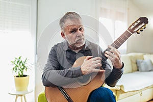 Mature man in casual clothes is smiling while playing guitar at home