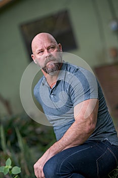 Handsome mature man sitting with arm on leg. Man is bald with graying beard