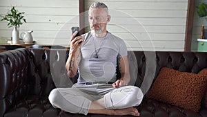 Handsome mature man sitting on the couch at home and communicating with somebody through video chat on his smartphone