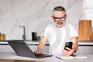 Handsome mature man talking on mobile phone while using laptop at the table at home