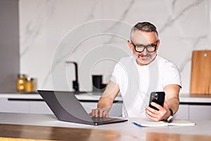 Handsome mature man talking on mobile phone while using laptop at the table at home