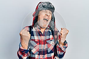 Handsome mature man wearing winter hat with ear flaps crazy and mad shouting and yelling with aggressive expression and arms