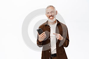 Handsome mature man with white beard, pointing finger at smartphone, showing mobile phone app, standing over white