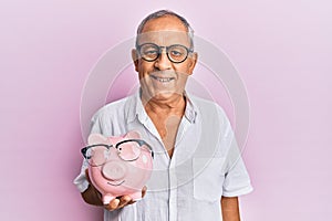 Handsome mature senior man holding piggy bank with glasses looking positive and happy standing and smiling with a confident smile