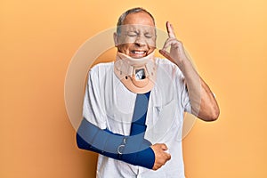 Handsome mature senior man wearing cervical collar and arm on sling gesturing finger crossed smiling with hope and eyes closed