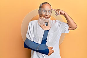 Handsome mature senior man wearing cervical collar and arm on sling smiling pointing to head with one finger, great idea or