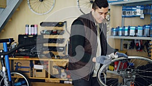 Handsome mechanic is cleaning bicycle wheel outside and inside, hub and spokes with piece of cloth and listening to