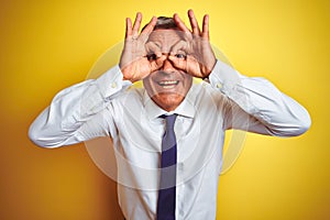 Handsome middle age businessman standing over isolated yellow background doing ok gesture like binoculars sticking tongue out,