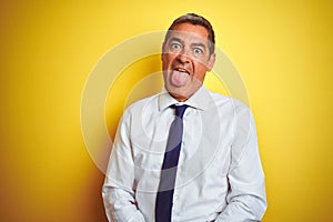 Handsome middle age businessman standing over isolated yellow background sticking tongue out happy with funny expression