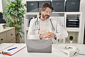 Handsome middle age doctor man working at the clinic checking the time on wrist watch, relaxed and confident