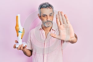 Handsome middle age man with grey hair holding anatomical model of knee joint with open hand doing stop sign with serious and
