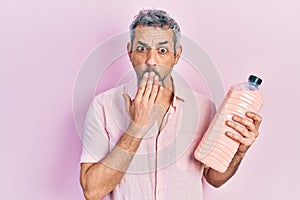 Handsome middle age man with grey hair holding detergent bottle covering mouth with hand, shocked and afraid for mistake