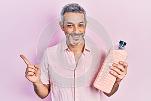 Handsome middle age man with grey hair holding detergent bottle smiling happy pointing with hand and finger to the side
