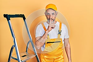 Handsome middle age man with grey hair holding ladder asking to be quiet with finger on lips