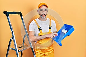 Handsome middle age man with grey hair holding roller painter making fish face with mouth and squinting eyes, crazy and comical