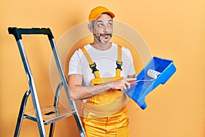 Handsome middle age man with grey hair holding roller painter smiling looking to the side and staring away thinking