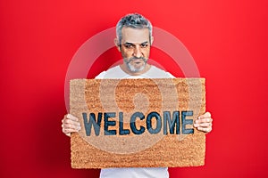 Handsome middle age man with grey hair holding welcome doormat skeptic and nervous, frowning upset because of problem