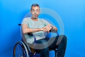 Handsome middle age man with grey hair sitting on wheelchair puffing cheeks with funny face