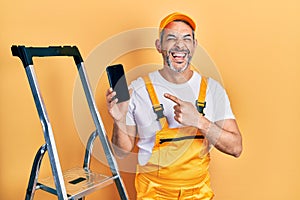 Handsome middle age man with grey hair standing by ladder showing smartphone smiling and laughing hard out loud because funny