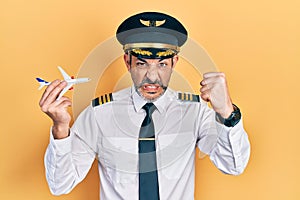 Handsome middle age man with grey hair wearing airplane pilot uniform holding toy plane annoyed and frustrated shouting with