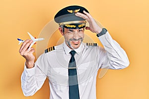 Handsome middle age man with grey hair wearing airplane pilot uniform holding toy plane stressed and frustrated with hand on head,