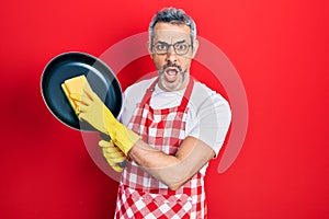 Handsome middle age man with grey hair wearing apron holding scourer washing pan afraid and shocked with surprise and amazed