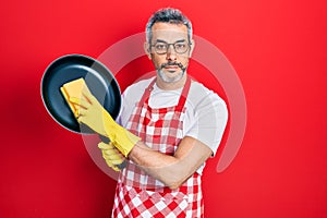Handsome middle age man with grey hair wearing apron holding scourer washing pan relaxed with serious expression on face