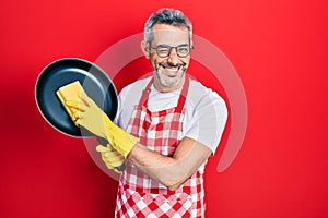 Handsome middle age man with grey hair wearing apron holding scourer washing pan winking looking at the camera with sexy
