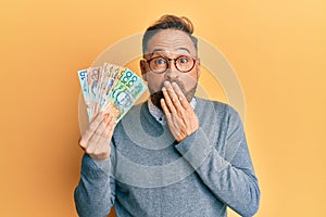 Handsome middle age man holding australian dollars covering mouth with hand, shocked and afraid for mistake