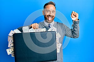 Handsome middle age man holding briefcase full of dollars and handcuffs sticking tongue out happy with funny expression