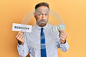 Handsome middle age man holding economy message and holding 1 dollar in shock face, looking skeptical and sarcastic, surprised