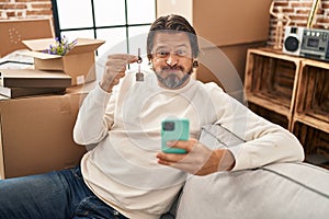 Handsome middle age man holding keys of new home doing video call puffing cheeks with funny face