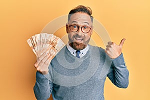 Handsome middle age man holding south african 20 rand banknotes pointing thumb up to the side smiling happy with open mouth