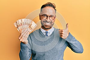 Handsome middle age man holding south african 20 rand banknotes smiling happy and positive, thumb up doing excellent and approval