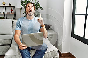 Handsome middle age man using computer laptop on the sofa amazed and surprised looking up and pointing with fingers and raised