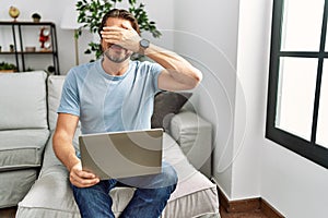 Handsome middle age man using computer laptop on the sofa smiling and laughing with hand on face covering eyes for surprise