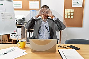 Handsome middle age man wearing call center agent headset at the office doing ok gesture like binoculars sticking tongue out, eyes