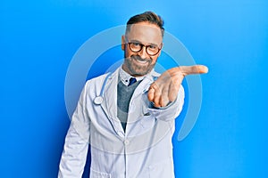 Handsome middle age man wearing doctor uniform and stethoscope smiling friendly offering handshake as greeting and welcoming