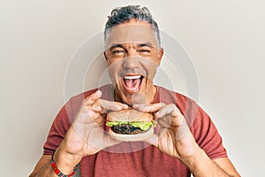 Handsome middle age mature man eating a tasty classic burger smiling and laughing hard out loud because funny crazy joke