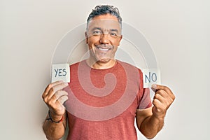 Handsome middle age mature man holding yes and no reminder smiling with a happy and cool smile on face