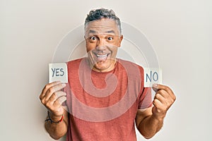 Handsome middle age mature man holding yes and no reminder smiling and laughing hard out loud because funny crazy joke