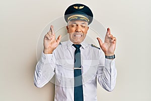 Handsome middle age mature man wearing airplane pilot uniform gesturing finger crossed smiling with hope and eyes closed