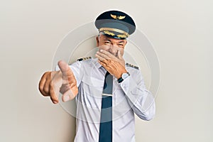 Handsome middle age mature man wearing airplane pilot uniform laughing at you, pointing finger to the camera with hand over mouth,