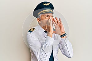 Handsome middle age mature man wearing airplane pilot uniform shouting angry out loud with hands over mouth