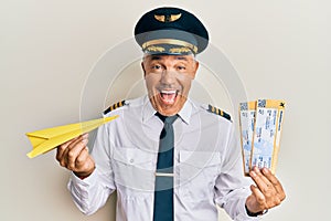 Handsome middle age mature pilot man holding paper plane and boarding pass smiling and laughing hard out loud because funny crazy