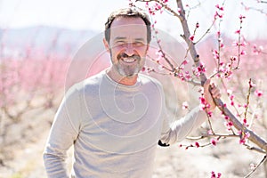 Handsome middle age senior man smiling cheerful on a peach trees garden with pink petals on a sunny day of spring